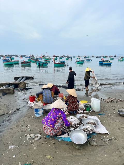      People sorting fish on a beach, boats in the background.
  