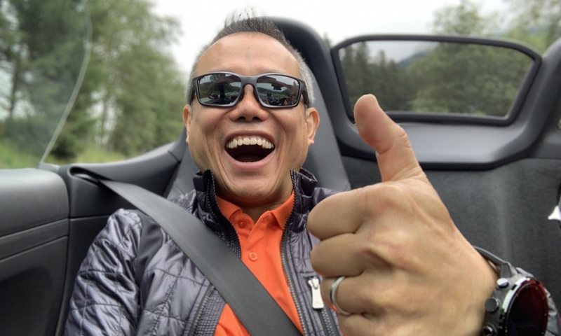 A man smiling and giving a thumbs up while wearing sunglasses in a convertible.