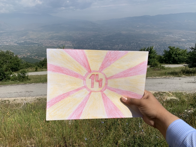       A hand holding a drawing with a scenic landscape in the background.
  