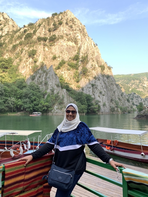       A person smiling in front of a scenic rocky lake.
  