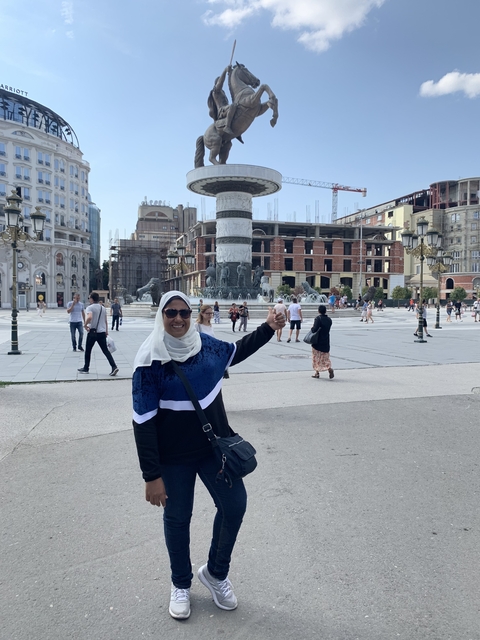       Woman in front of a fountain with surrounding buildings.
  