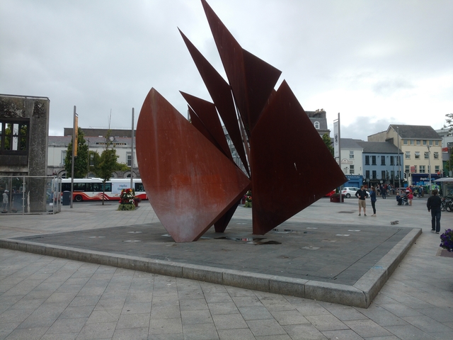 A large public plaza with a modern sculpture and people around.