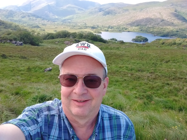 A man taking a selfie with a lush green landscape and a lake in the background.