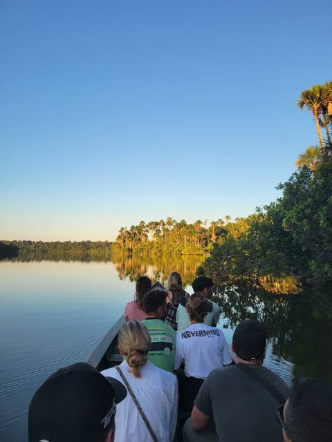       People in a boat on a calm river with a reflection of trees.
  
