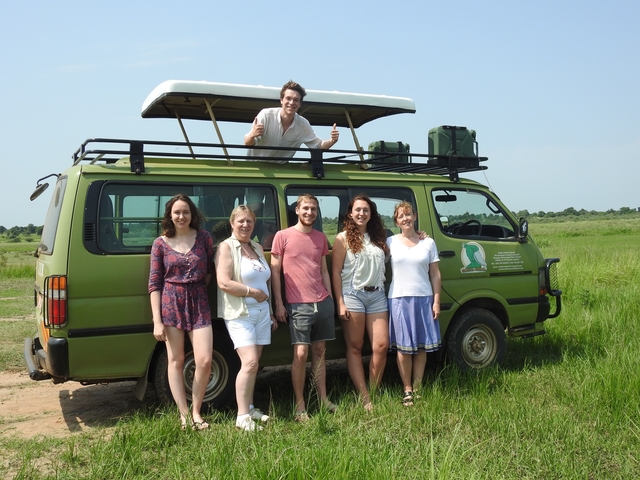 Group of travelers posing with a safari vehicle in a grassy area.