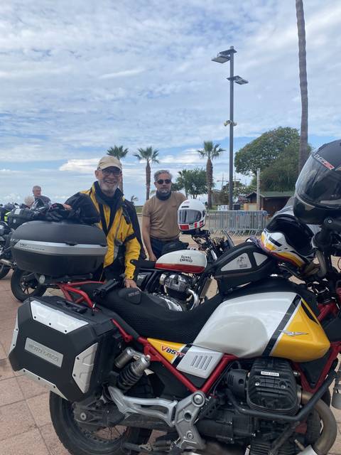       Two people with motorcycles, palms, and cloudy sky.
  