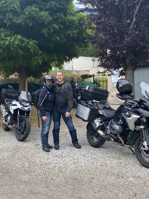       Two people posing with motorcycles in a natural setting.
  