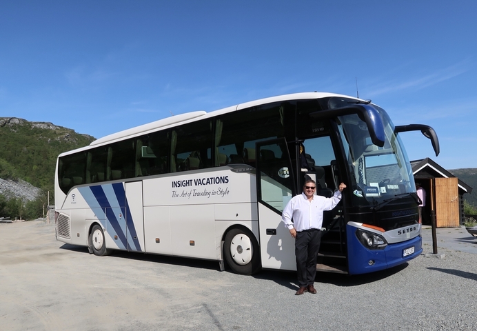 A person posing beside a large tour bus with mountains in the background.