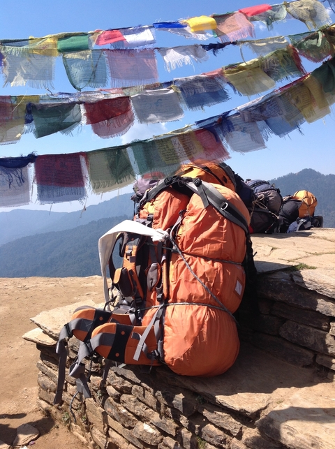       Backpacks resting on a ledge with prayer flags above and mountains beyond.
  