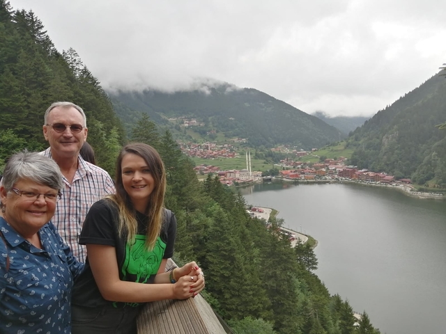 A family posing with a view of lush green hills and a village by a lake.