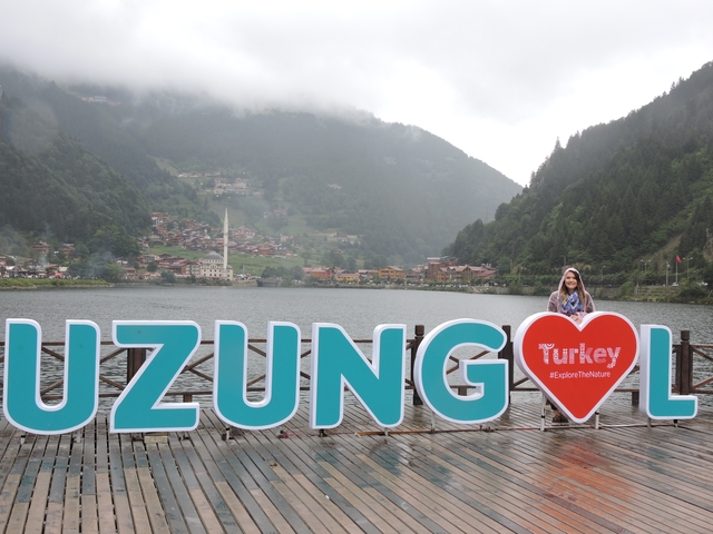 A person posing next to a large Uzungöl sign with a village and nature backdrop.