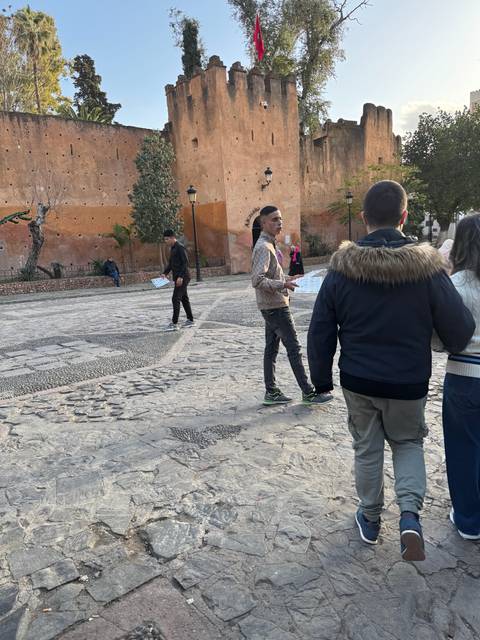People walking in front of historic walls and a Moroccan flag.