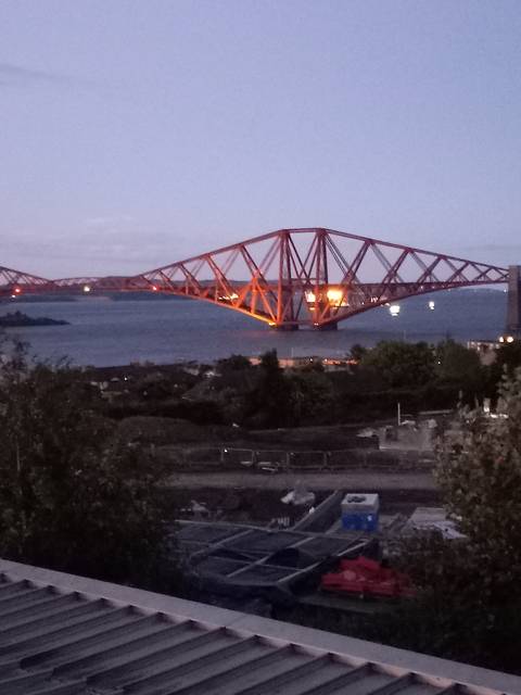 Forth Bridge at dusk with bright lights against the darkening sky.