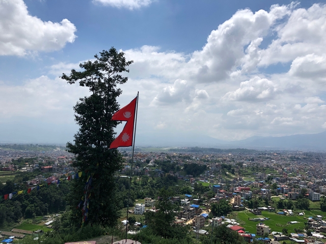       Nepal flag on a pole with panoramic city view and mountains.
  