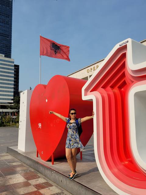       Person posing with a large red sculpture and an Albanian flag.
  