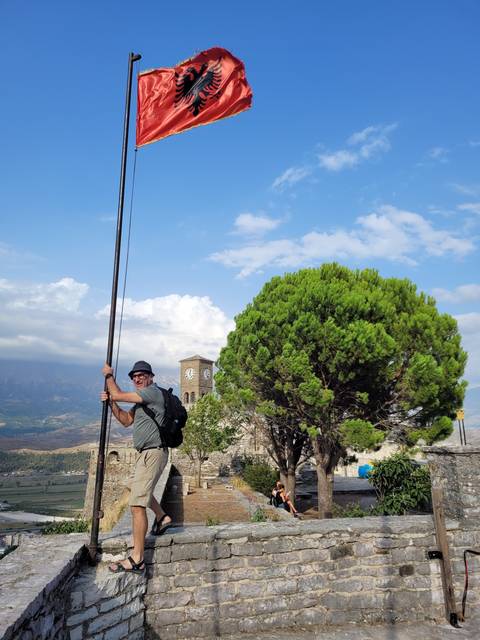       Man posing next to an Albanian flag with a historical building in the background.
  