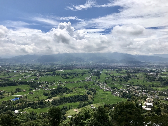 Panoramic view of lush green fields with mountains and cloudy sky in the background.