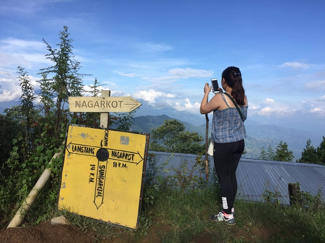 Person taking a photo near a sign pointing to Nagarkot with mountains in the background.