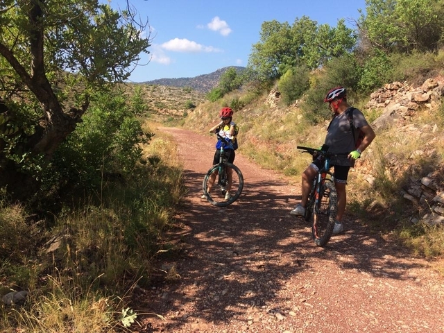 Two cyclists wearing helmets on a dirt path surrounded by hills.
