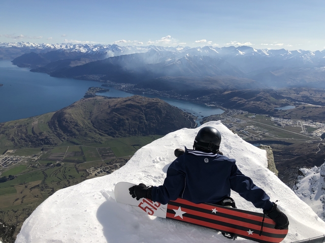 Person sitting on snowy mountain peak overlooking a body of water.