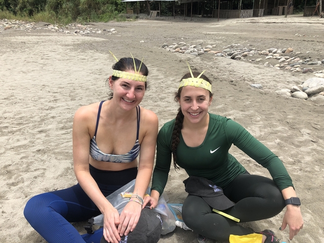      Two women seated on a sandy area wearing hats with reeds.
  
