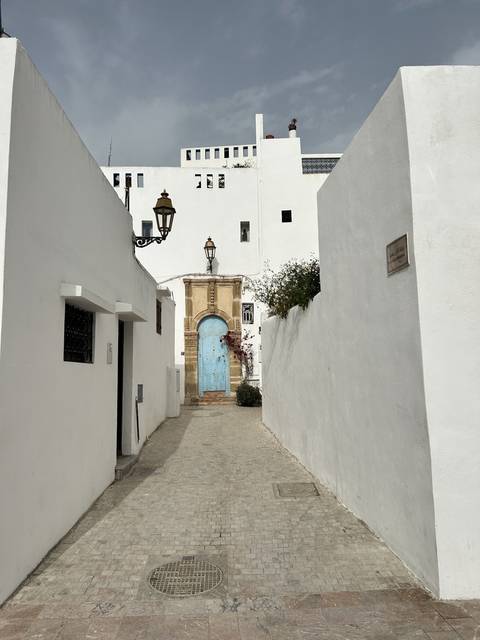       Narrow street with white buildings and a blue fountain.
  