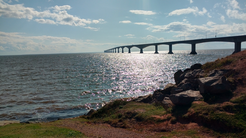       Long bridge over shimmering water with rocky shore.
  