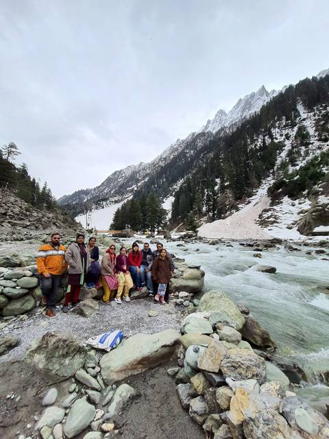 Group of people sitting on rocks by a river with snowy backdrop.