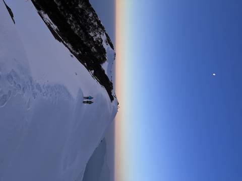       Two people standing on a snowy ridge with the moon visible in the sky.
  