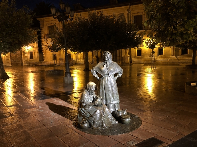       Sculpture of women in traditional attire on a wet street at night.
  