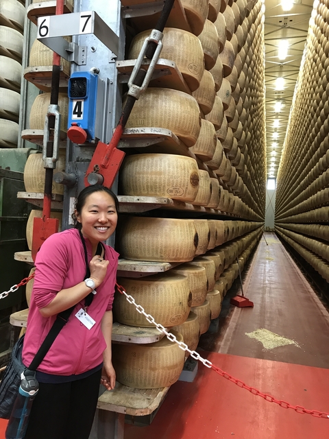 Woman posing next to large cheese wheels in a storage room.