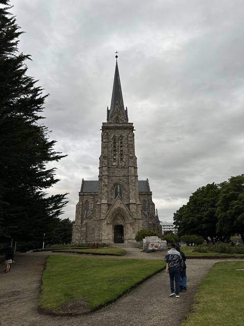       Tall stone church with a pointed spire and tourists walking nearby.
  