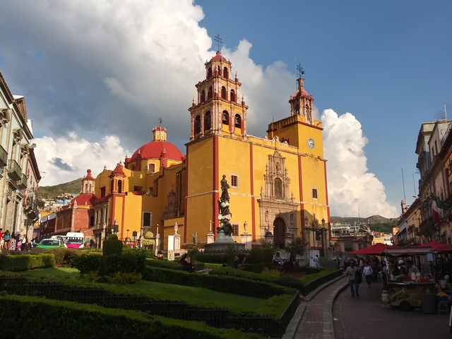       A brightly colored cathedral with a garden and cloudy sky.
  