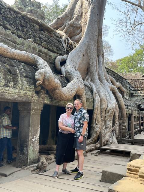       Two people standing beside a massive tree growing over ancient ruins.
  