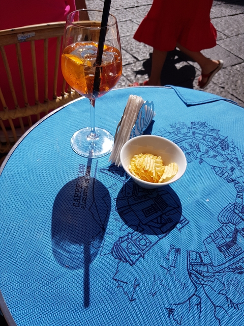       Outdoor cafe table with wine glass and snacks on a decorated blue tablecloth.
  