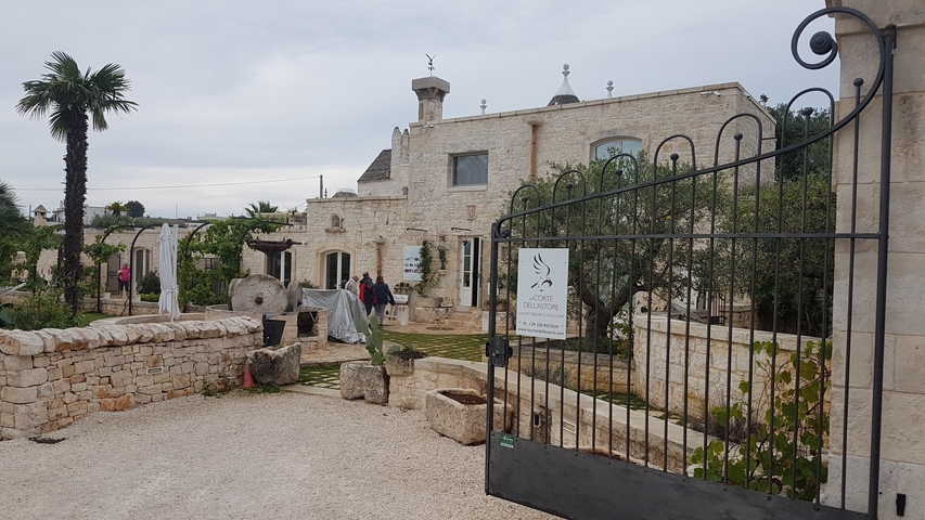       People near a stone building with gate and plants.
  