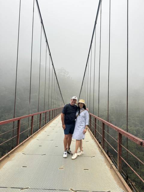       A couple standing on a suspension bridge in a misty setting.
  