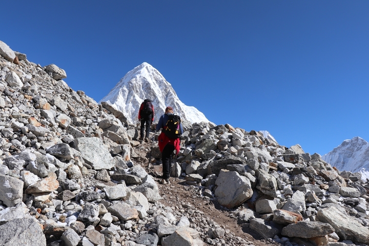 Two trekkers on a rocky path towards snow-capped peaks.
