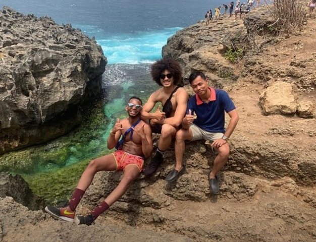 Three people smiling and giving thumbs up on rocky coastline with clear blue water.