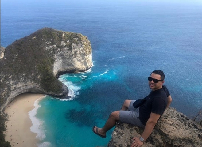 Person sitting on a cliff edge overlooking a beach and clear blue ocean.