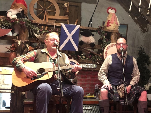       Two men singing and playing guitar in a rustic setting, Scottish flag in the background.
  