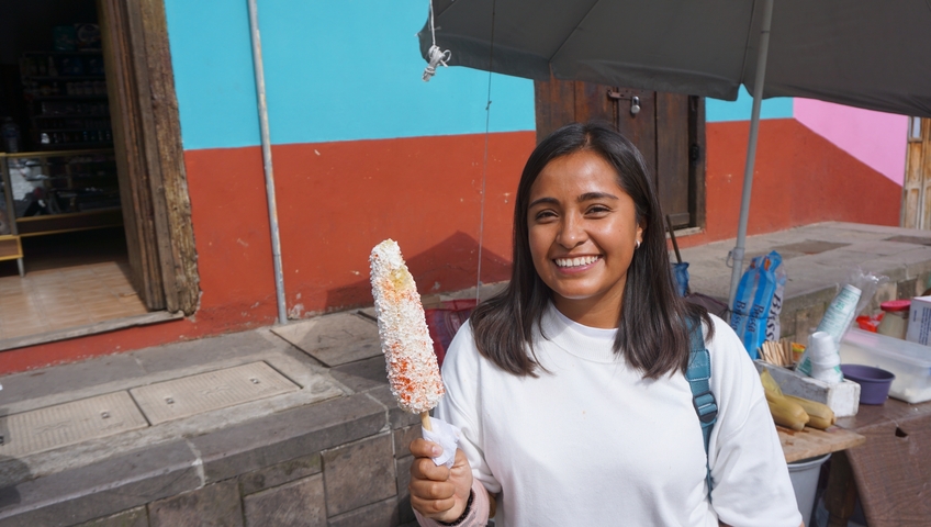       Smiling woman holding a snack on a colorful street.
  