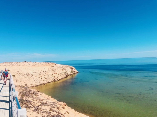 A person on a boardwalk overlooking a clear ocean and rocky coastline.