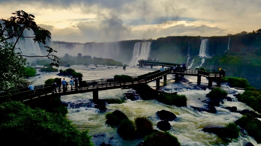 Tourists walking on a bridge over Iguazu Falls with mist in the background.