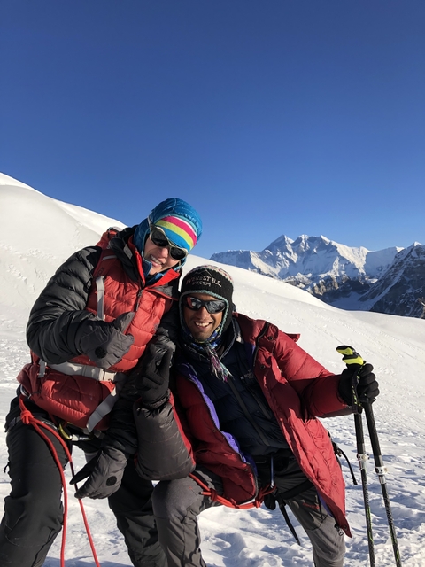       Two climbers posing on a snowy Himalayan slope, with mountains behind.
  