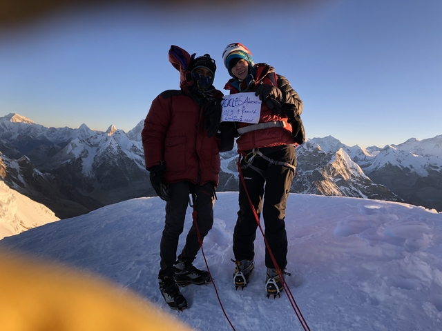       Two mountaineers on a snowy peak with mountains in the background.
  