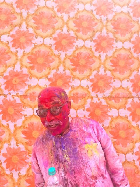       Person covered in Holi colors standing against a floral patterned wall.
  