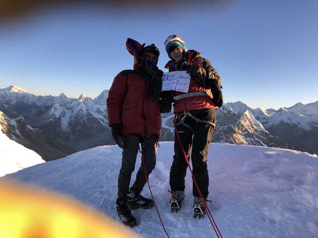 Two hikers holding a sign at the top of a snowy peak.