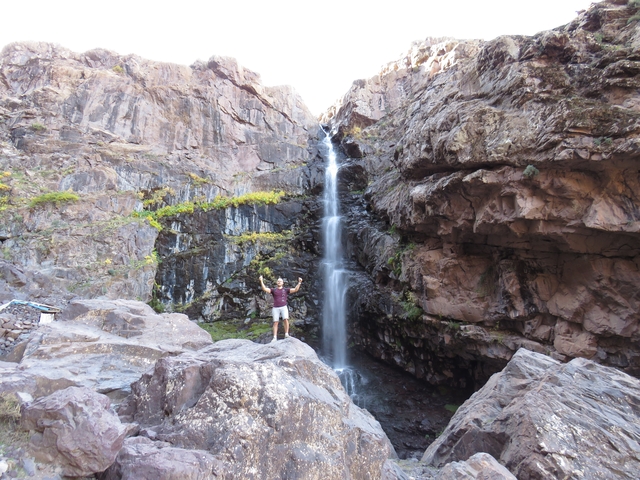 Person posing in front of a narrow waterfall cascading down a steep rocky cliff.