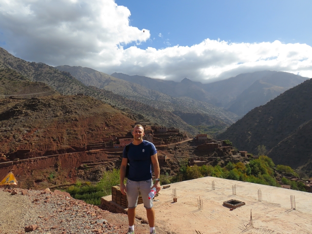 Man smiling with a scenic mountain village and lush landscape under a cloudy sky.